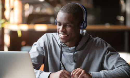 Smiling african young man student wear headphones looking at laptop screen writing notes, happy black guy e-learning in internet study online with skype teacher on computer watch webinar sit in cafe